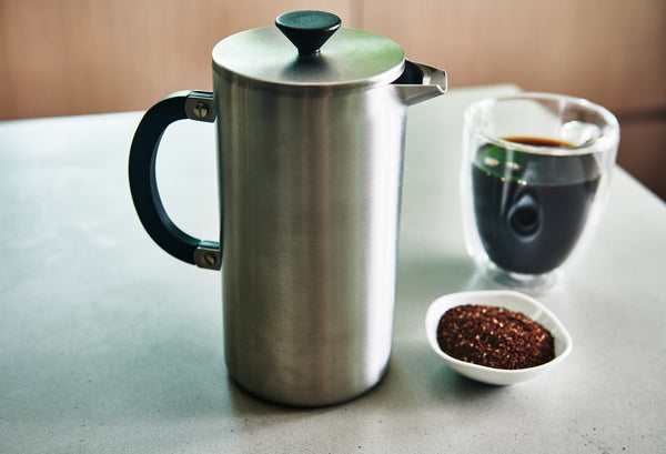 A stainless steel French press coffee maker with a black handle and lid knob sits on a light gray tabletop next to a glass of black coffee and a small dish of ground coffee beans.