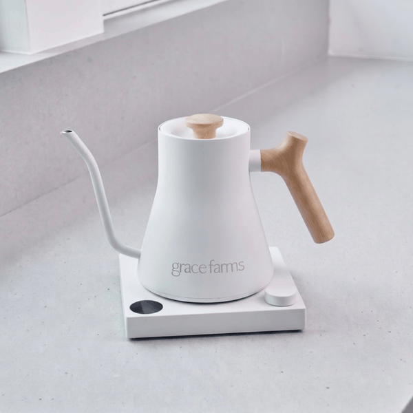 A white Grace Farms gooseneck kettle with a light wood handle and lid knob, sitting on a white electric kettle burner base on a light gray countertop.
