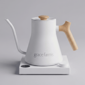 A white Grace Farms gooseneck kettle with a light wood handle and lid knob, resting on a white electric heating plate against a neutral gray background.