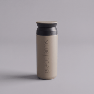 KINTO travel tumbler in sand beige, featuring the vertical white text 'grace farms' on the front, set against a solid gray background.