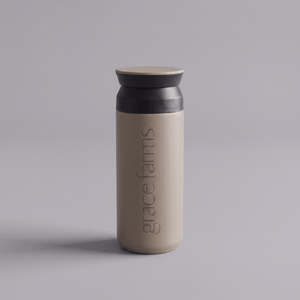 KINTO travel tumbler in sand beige, featuring the vertical white text 'grace farms' on the front, set against a solid gray background.