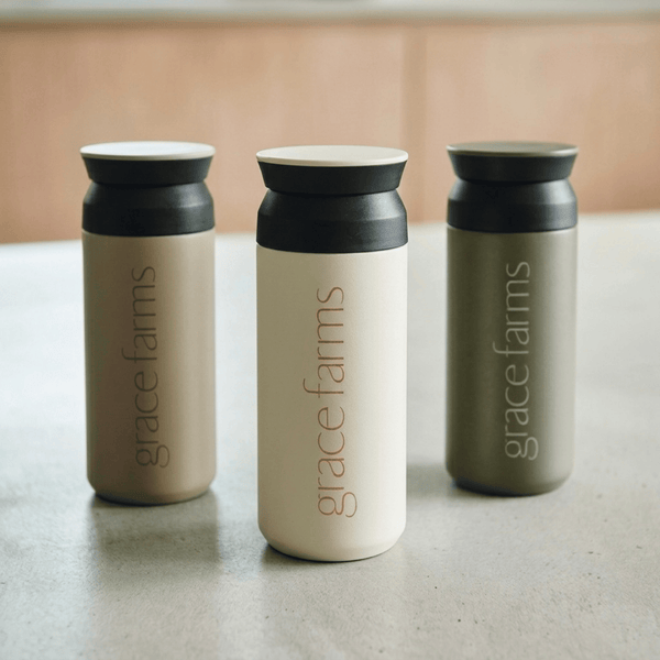 Three Grace Farms insulated tumblers with lids, shown in taupe, cream, and sage green on a countertop.